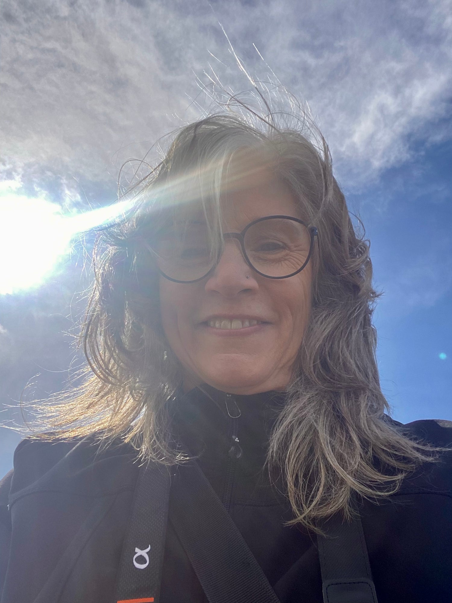 Person with glasses and a dark jacket against a blue sky with clouds