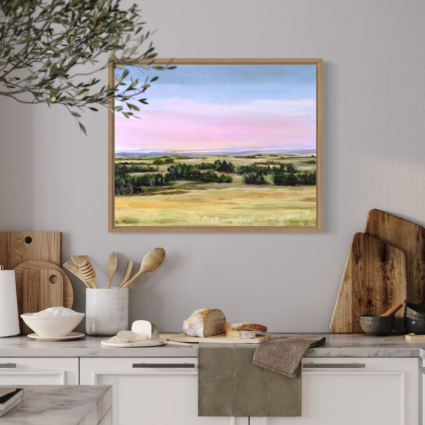 Framed landscape painting above a kitchen counter with wooden cutting boards and utensils.