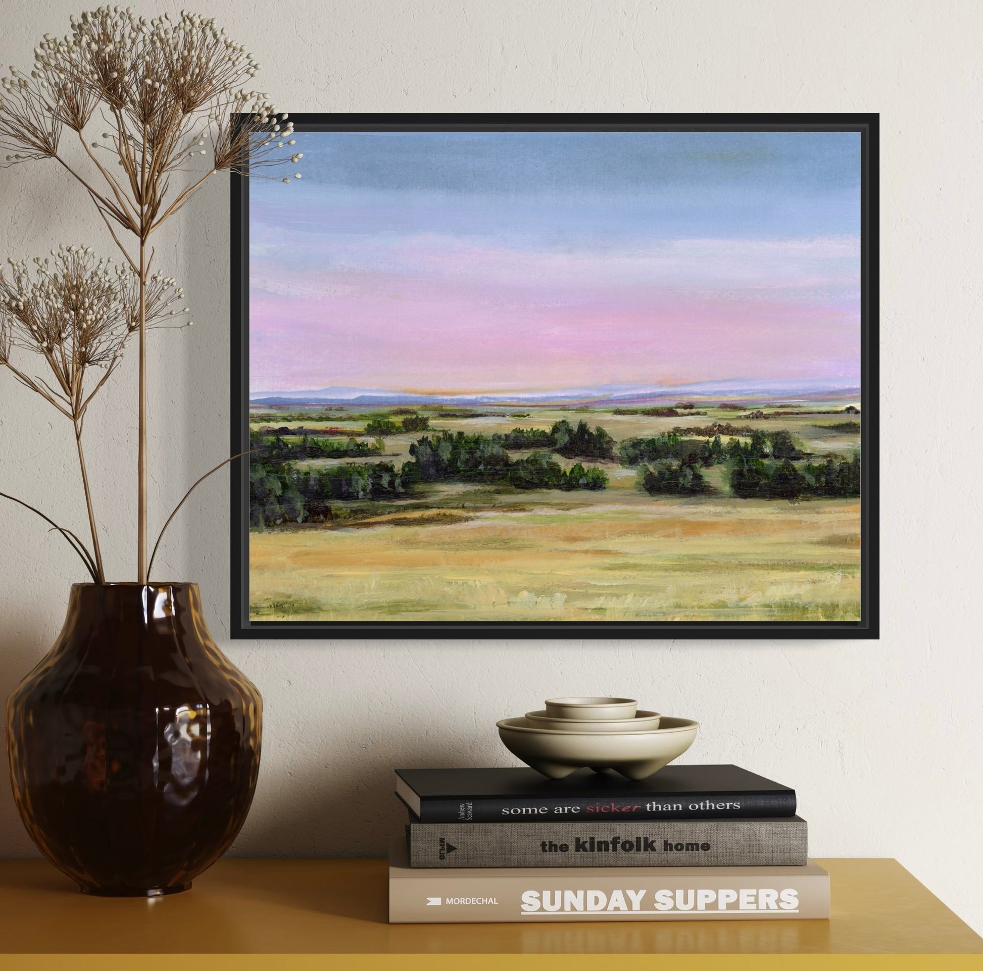 Framed landscape painting on a wall with a vase and books on a table below.