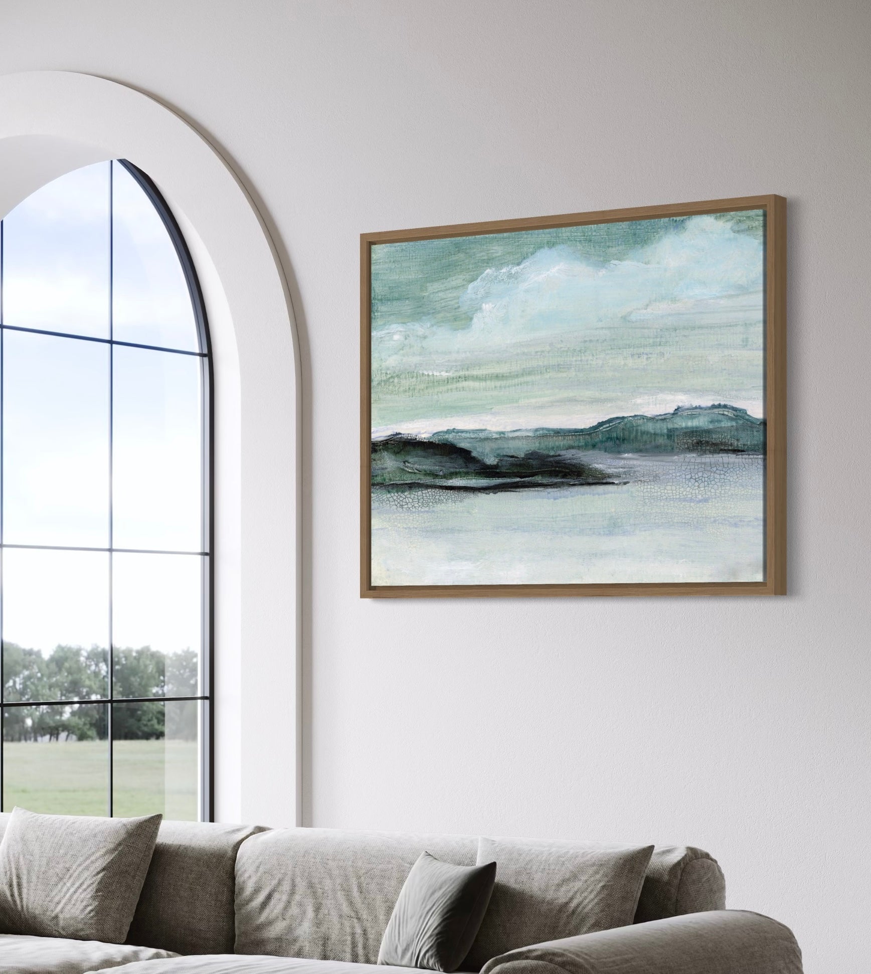 Framed landscape painting on a wall above a sofa with large windows in the background