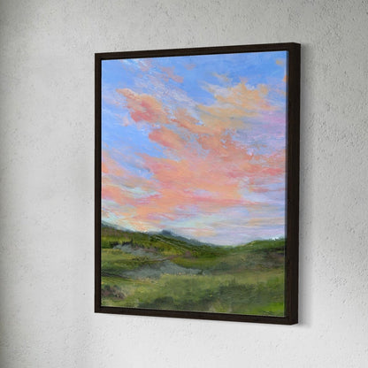 Framed painting of a landscape with pink clouds and green fields on a white wall.